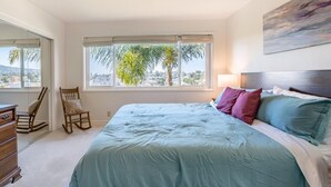 A bedroom with a large bed, a rocking chair, a dresser, and a window with a city view. - Spacious Condo with Stunning View (Oakland)