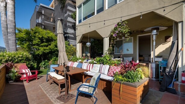 A patio with a wooden table, chairs, and a built-in bench with cushions. There are potted plants and a pergola with string lights. - Spacious Condo with Stunning View (Oakland)