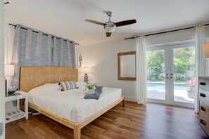A bedroom with a wooden bed, a ceiling fan, a bedside table with a lamp, and a view of a swimming pool through a glass door. - Private Palm Beach Pool Home Putting Green near BEACH Restaurants (North Palm Beach)