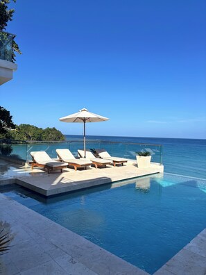 An infinity pool with lounge chairs and an umbrella, overlooking a clear blue sea. - skyfall perfectly positioned on beachfront with panoramic views of the sanctuary (Ocho Rios)