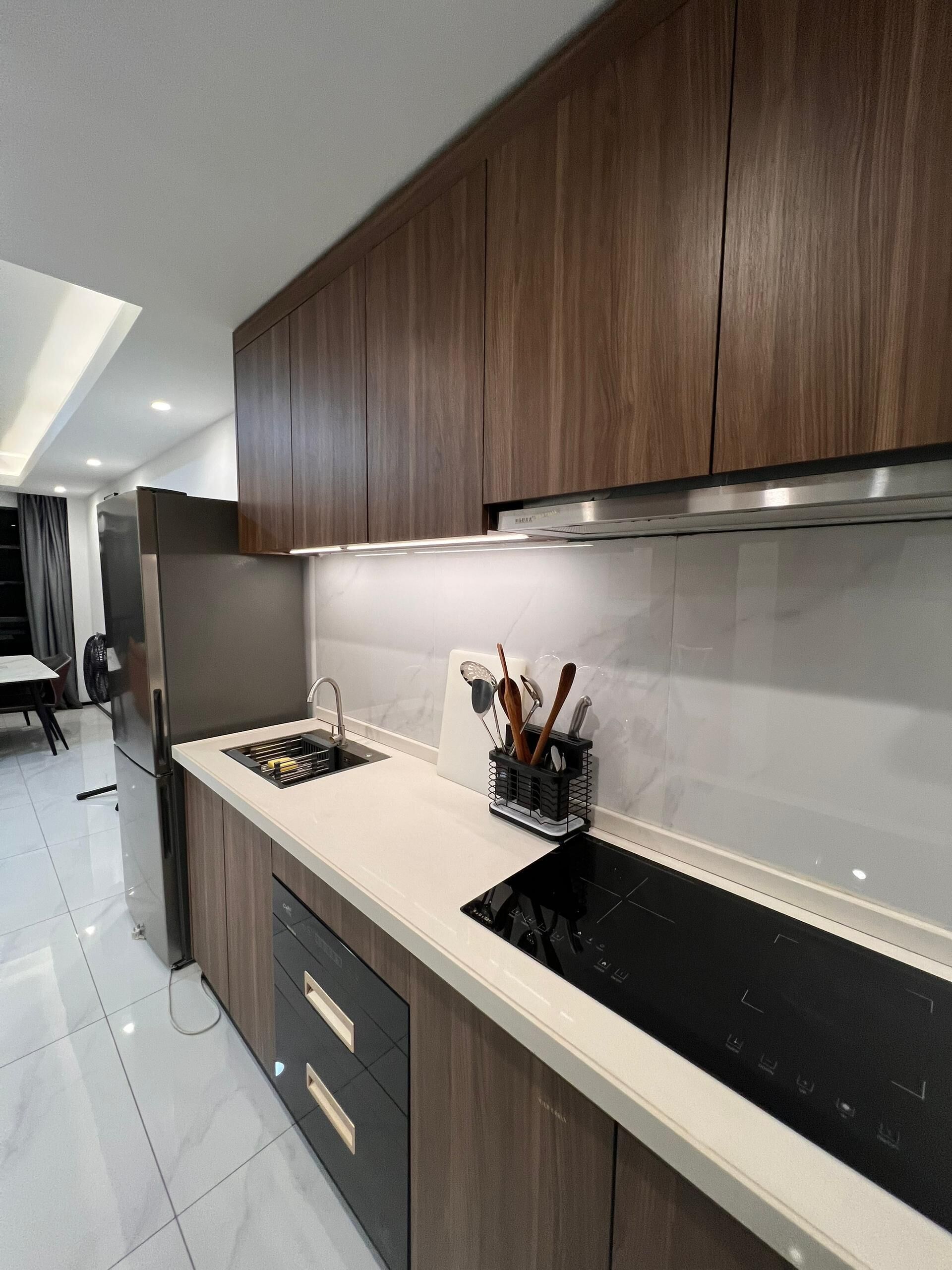 Eine moderne Küche mit dunklen Holzschränken, einem Edelstahlkühlschrank, einer schwarzen Induktionskochplatte und einer weißen Arbeitsplatte.