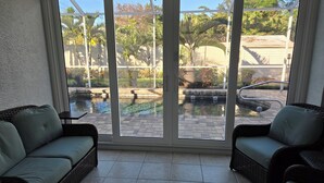 Una stanza con una porta scorrevole in vetro che dà su un giardino con piscina, due divani in vimini e un pavimento piastrellato.