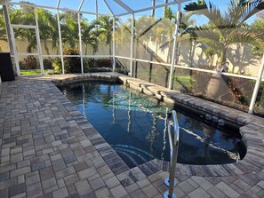 Una piscina con borde de ladrillo, rodeada por un cerramiento de vidrio donde se ven palmeras y plantas.