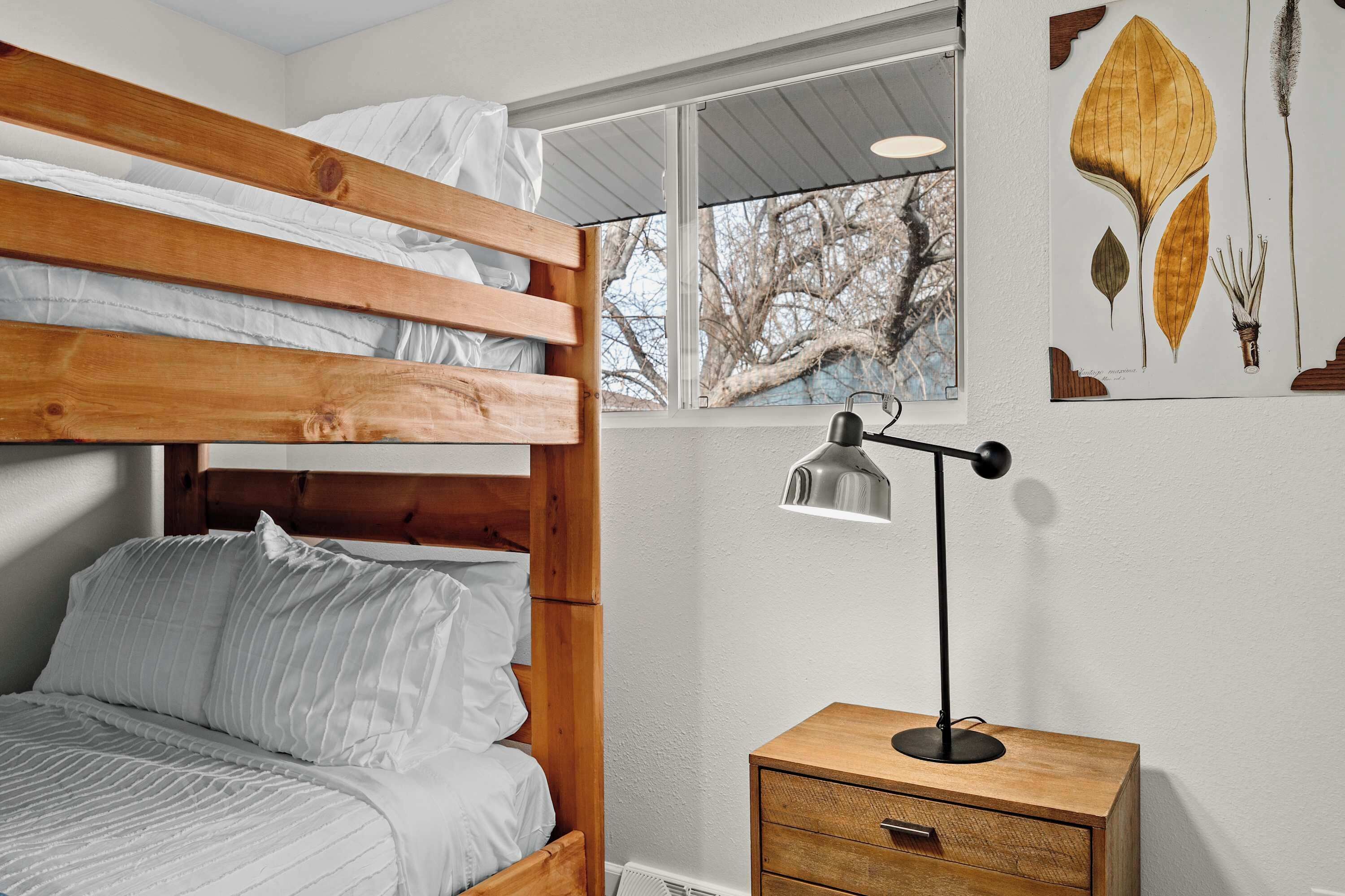 Una cama individual con ropa de cama blanca, una mesita de noche con lámpara y una ventana con vistas a árboles.