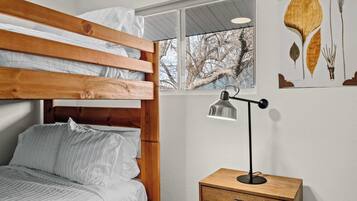 Una cama individual con ropa de cama blanca, una mesita de noche con lámpara y una ventana con vistas a árboles.