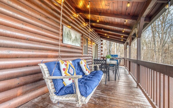 A wooden log cabin with a porch, a swing seat with blue cushions, and a table with chairs. - 2MstrSuites~Pickleball~Golf~Hottub~Firepit~Gameroom~pooltable~Views~Sleeps 16 (Talking Rock)