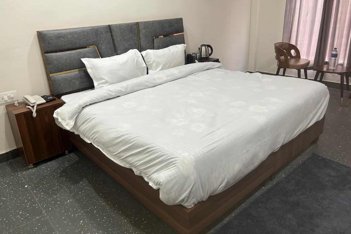 A neatly made bed with a grey headboard, a bedside table with a phone and a kettle, and a chair in the corner.