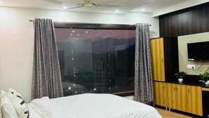 Une chambre moderne avec un grand lit, un ventilateur de plafond, une télévision et une vue sur le paysage urbain par la fenêtre.