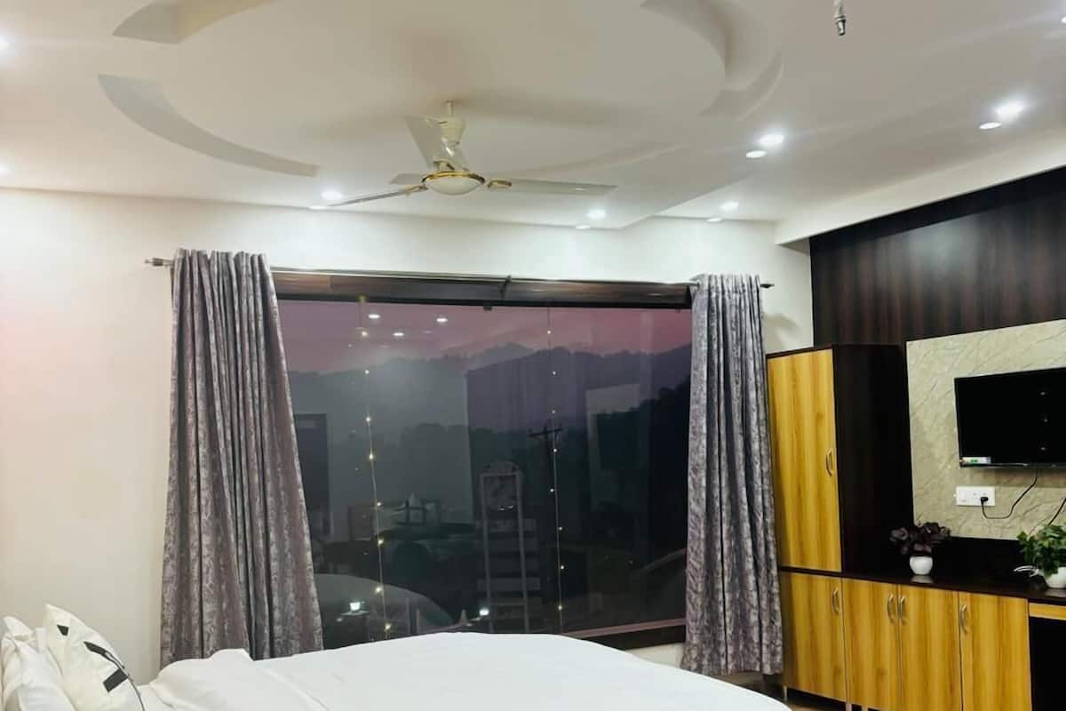 A modern bedroom with a large bed, a ceiling fan, a TV, and a view of the cityscape through the window.