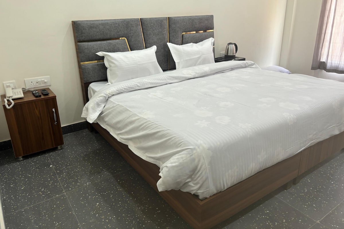 A neatly made bed with a grey upholstered headboard, white bedding, and a bedside table with a phone and remote controls.