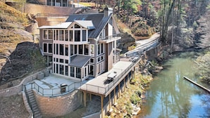Een meerlaags huis met een terras met uitzicht op een rivier, omgeven door bomen en een rotsachtig landschap.
