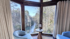 Een kamer met een groot raam met uitzicht op een bos en een rivier. Er staan twee stoelen en een klein tafeltje met een lamp.