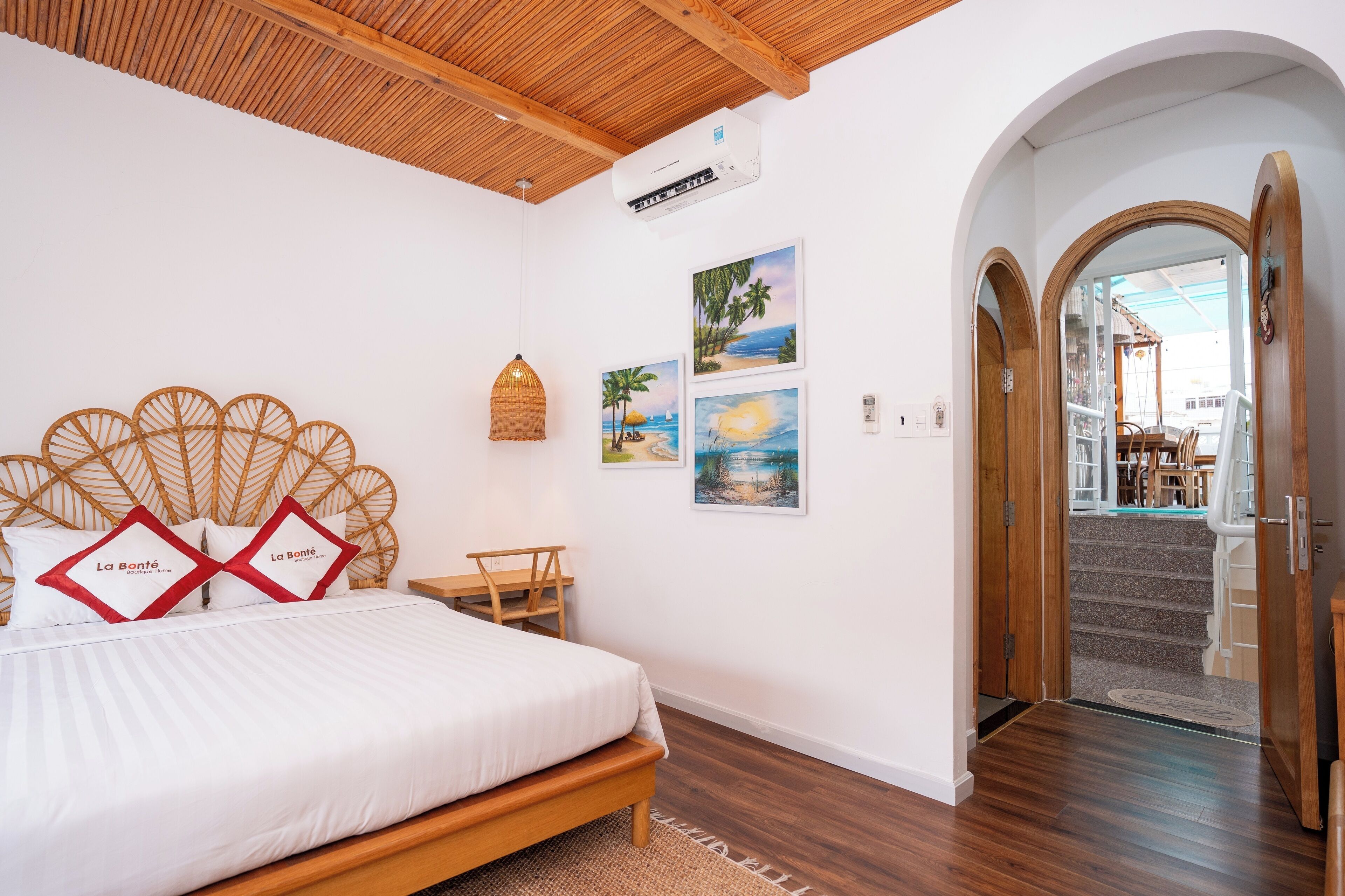 A bedroom with a wooden headboard, a bed with white bedding, a wooden bench, and a small table. There are three framed pictures on the wall, and an air conditioning unit above the bed.