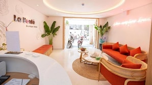 A reception area with a white reception desk, a wicker sofa with orange cushions, and a round table with a small plant. - La Bonté Boutique Home Vung Tau (Ho Chi Minh City)