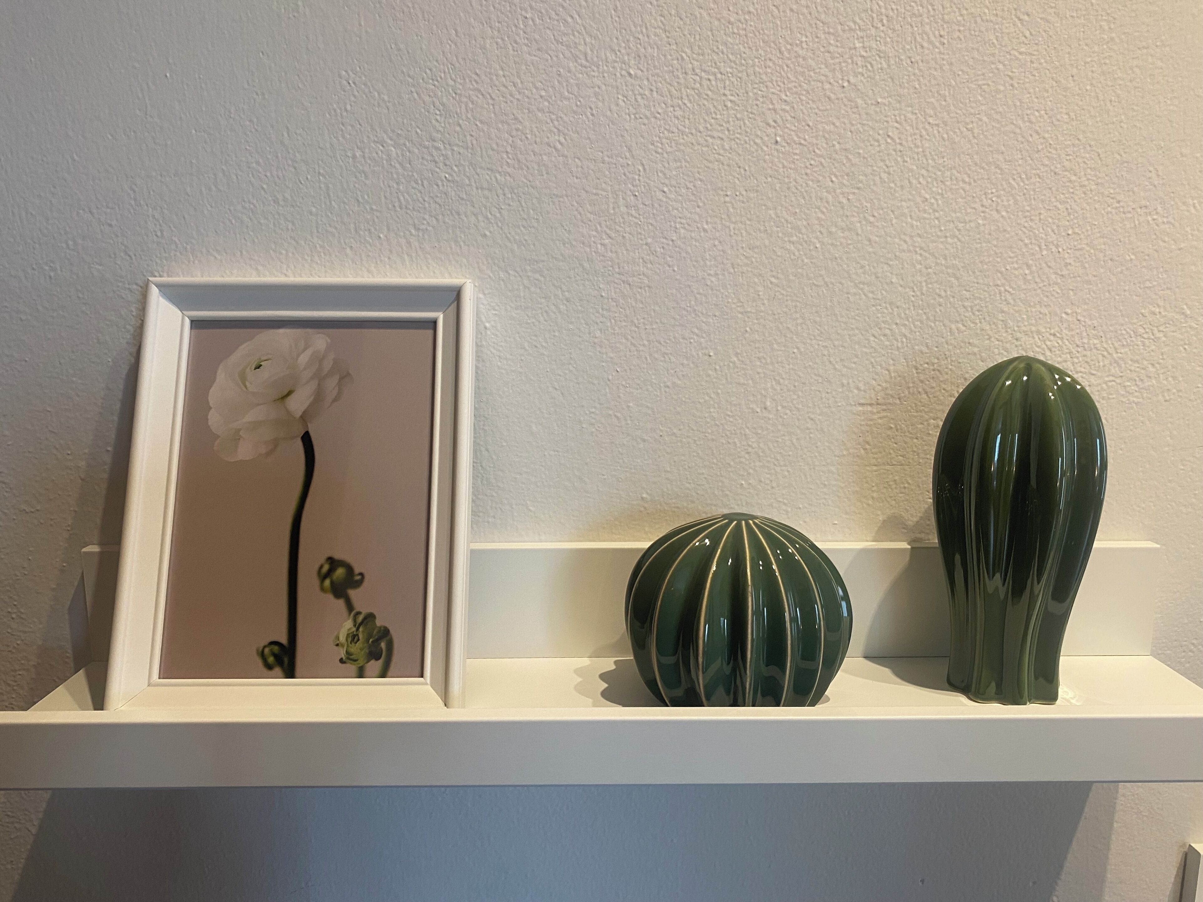 En hvid reol med et indrammet billede af en blomst, en grøn vase og en grøn keramikskål.