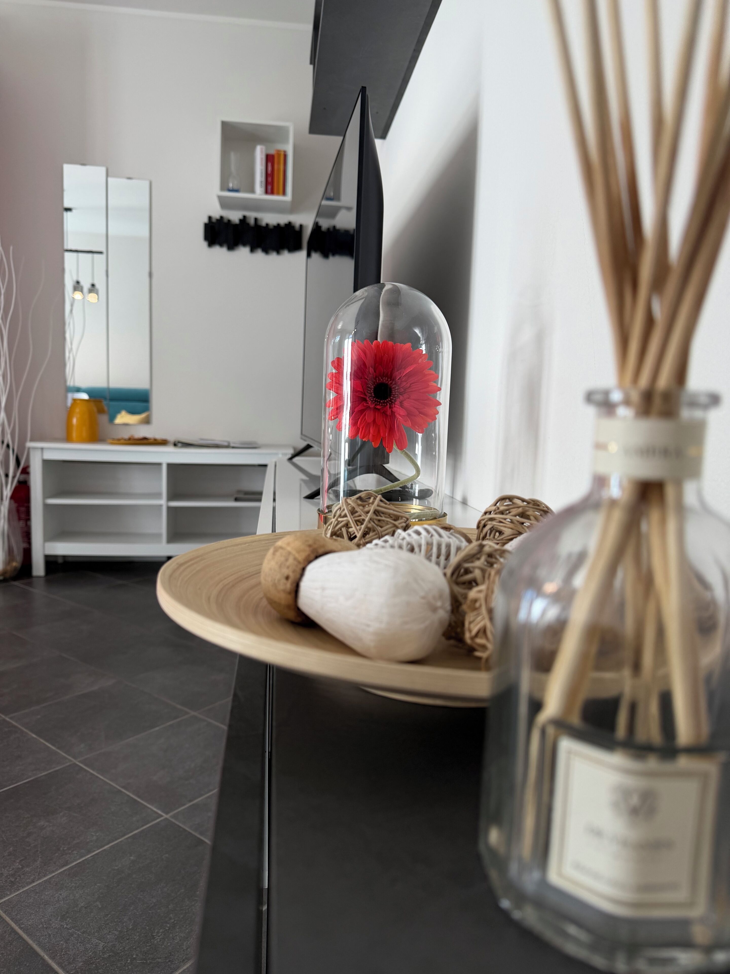 Un salon moderne doté d’une verrière où est exposée une fleur rouge, d’un diffuseur de parfum à bâtonnets et d’une table ronde en bois agrémentée d’objets décoratifs.