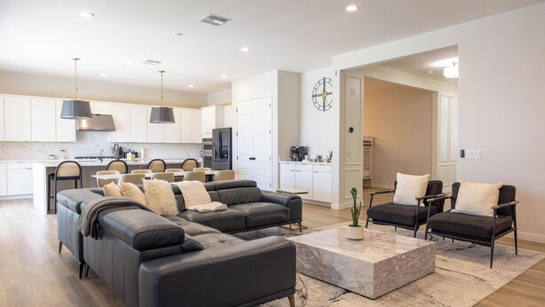 A modern living room with a sectional sofa, a coffee table, and two armchairs. A kitchen with white cabinets and a dining area with a table and chairs is visible in the background. - New Modern Luxury 8-bed mins to AAG, Fwy, Clubhouse. Ideal Sports or Ext Stay  (Mesa)