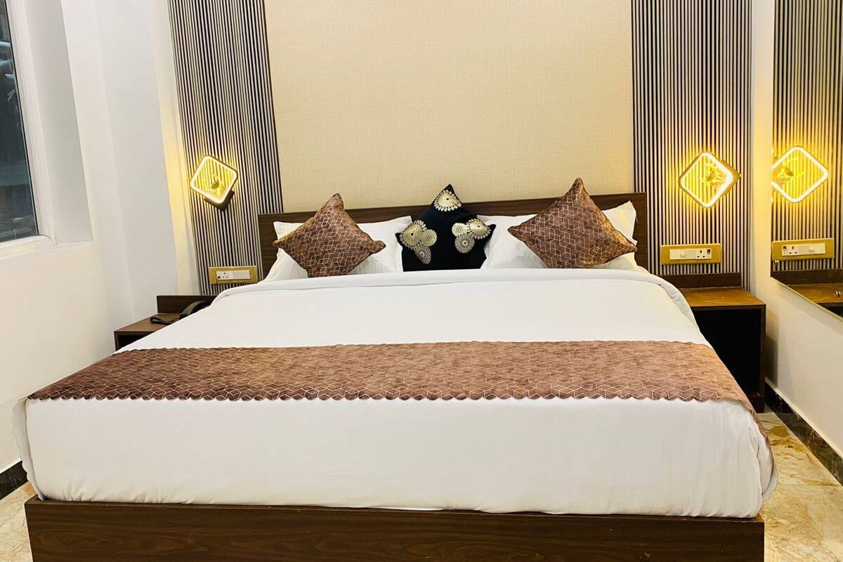 Una cama bien hecha con una manta marrón, un cabecero de madera y almohadones decorativos. Se observan mesitas de noche con lámparas y un aplique en la pared.