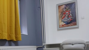 Una habitación con una mesa blanca, una silla, un radiador, una cortina amarilla y un cuadro en la pared.