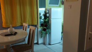 Un comedor con una mesa, sillas y una alfombra. Se ve un refrigerador y un televisor al fondo.