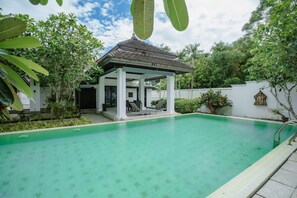 A swimming pool with a covered seating area, surrounded by lush greenery and a white wall with a decorative relief. - Number Nine Pool Villa  (Pa Klok)