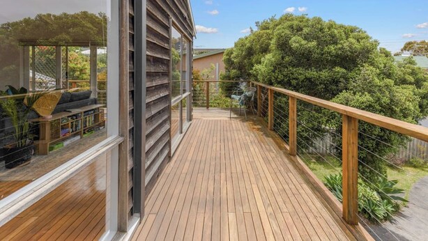 A wooden deck with a glass balustrade, a seating area with cushions, and a view of a green landscape. - The Balcony - pet friendly! (Jan Juc)