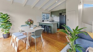 A modern kitchen with a central island, a dining area with a table and chairs, and a living area with a sofa and plants. - The Balcony - pet friendly! (Jan Juc)