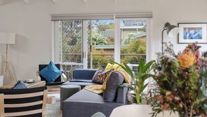 A modern living room with a grey sofa, a wooden chair, and a small table. There is a large window with a view of greenery outside. - The Balcony - pet friendly! (Jan Juc)