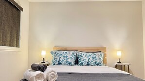 Ein ordentlich bezogenes Bett mit grauer Tagesdecke, zwei Kissen mit Blättermotiv und zwei Nachttischlampen.