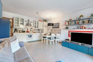 Μια σύγχρονη κουζίνα με λευκά ντουλάπια, μια τραπεζαρία με ένα λευκό τραπέζι και καρέκλες, μια μεγάλη τηλεόραση επίπεδης οθόνης και μια βιβλιοθήκη γεμάτη με βιβλία και διακοσμητικά αντικείμενα.