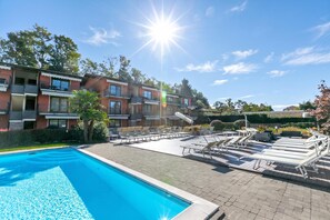 A swimming pool with lounge chairs and a sunshade, surrounded by a brick building with balconies. - Blu Oltrelago: Leggiuno Lake Maggiore Apt w/ Shared Pool & Garden (Leggiuno)