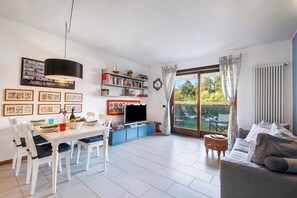A spacious living area with a dining table, chairs, a sofa, a TV, and a bookshelf. - Blu Oltrelago: Leggiuno Lake Maggiore Apt w/ Shared Pool & Garden (Leggiuno)