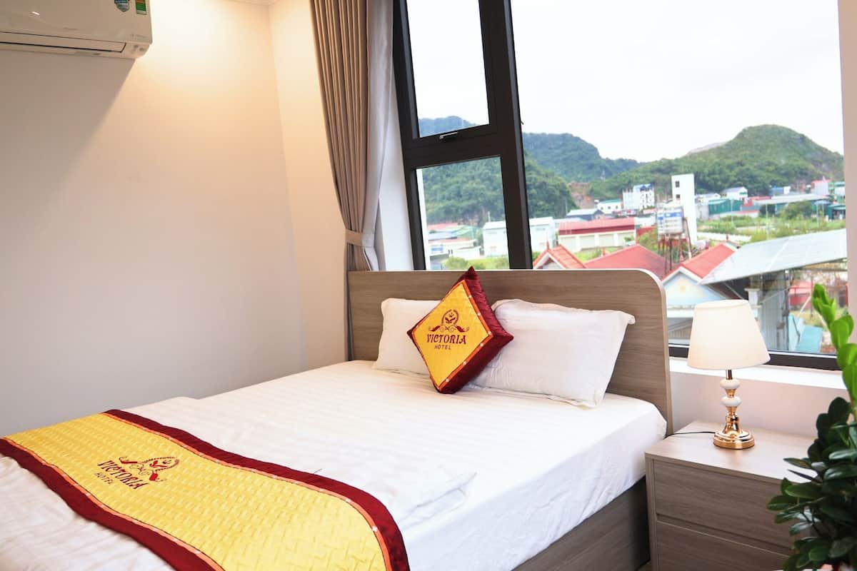 Ein Hotelzimmer mit Bett, Nachttisch, Lampe und Fenster mit Blick auf Gebäude und Hügel.