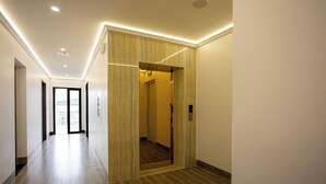 Un couloir moderne avec un plancher en bois, une porte d’ascenseur en verre et une vue sur l’extérieur grâce à une porte vitrée au fond.