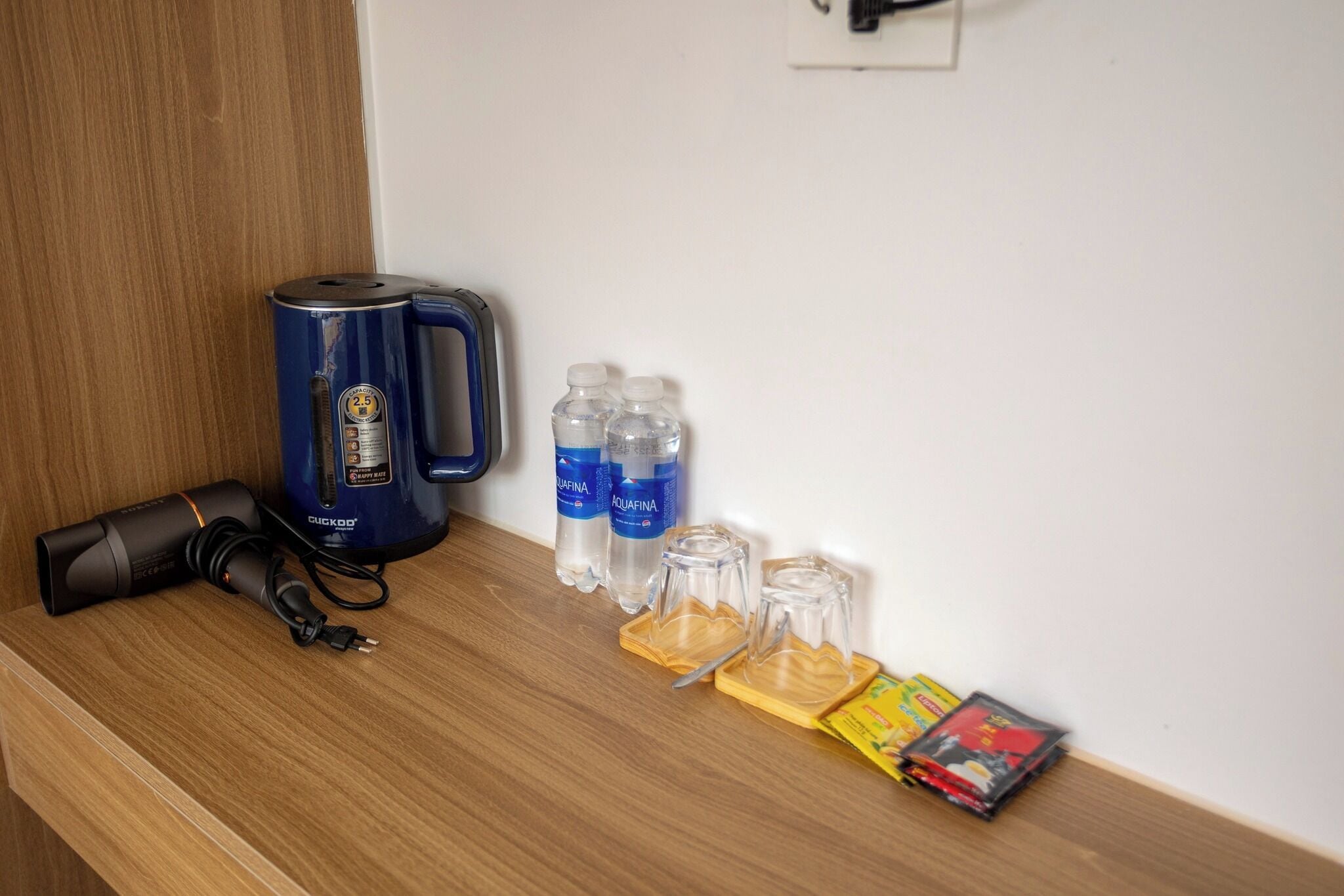 Une table en bois sur laquelle se trouvent une bouilloire électrique bleue, deux bouteilles d’eau, deux carafes en verre et quelques magazines.