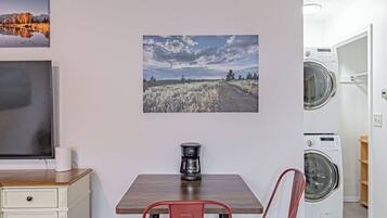 Una habitación pequeña con una mesa de madera, dos sillas rojas, cafetera, televisor y lavadora.