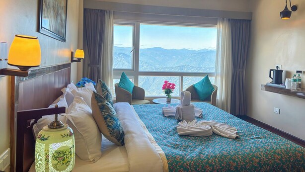 A hotel room with a large bed, a view of mountains, a desk with a lamp, and a shelf with toiletries. - Rare Himalayas Mapham (Darjeeling)