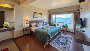 A hotel room with a large bed, a nightstand, a chair, a window with a view, and a wall-mounted TV. - Rare Himalayas Mapham (Darjeeling)