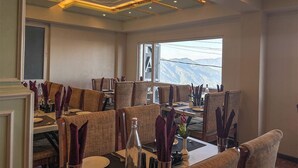 A dining area with multiple tables and chairs, a large window with a mountain view, and a modern ceiling design. - Rare Himalayas Mapham (Darjeeling)