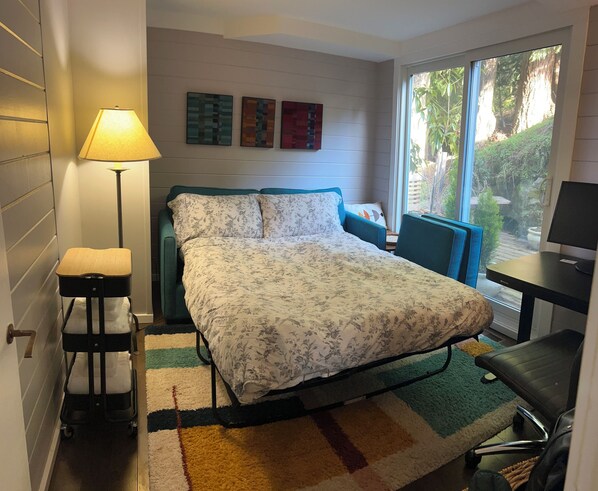 A bedroom with a bed, a desk, a chair, a lamp, and a window with a view of greenery. - Treehouse on Bowen - Sunsets + Patios near Beaches and Trails (Bowen Island)