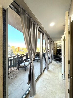 Varanda com cortinas cinzentas, vista para o horizonte urbano e área de estar com um banco e uma pequena mesa redonda.