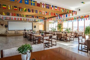 Un salón de conferencias con varias mesas y sillas, una pantalla de proyector y un ventanal con vistas. Hay banderas decorando las paredes.