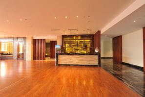 Zona de recepción de un hotel con piso de madera, mostrador de recepción con lámpara de diseño geométrico y un amplio ventanal al fondo.