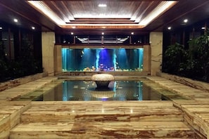 Amplia zona de piscina interior con una fuente central, rodeada de suelo y paredes de mármol, y un techo con iluminación empotrada.