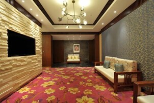 Habitación de hotel con alfombra roja, sofá, televisor montado en una pared texturizada y candelabro.