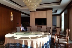 Una mesa de comedor redonda con tapa de vidrio, rodeada de sillas, en una habitación con una lámpara de araña grande, un televisor de pantalla plana y un ventanal amplio.