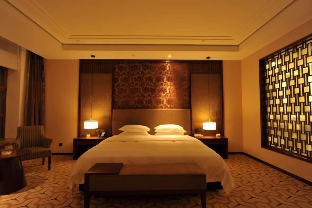 Een hotelkamer met een groot bed, twee nachtkastjes met lampen, een stoel, een bankje en een decoratief wandpaneel.