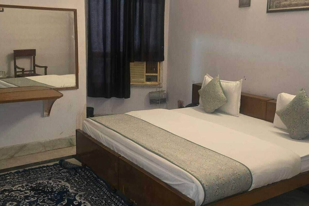 A bedroom with a bed, a nightstand, a mirror, a window with curtains, and a patterned rug.