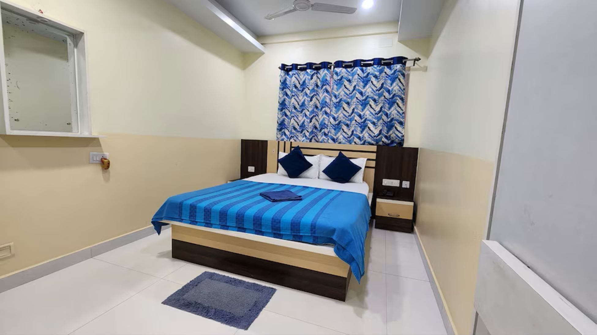 A bedroom with a bed, a nightstand, a wall-mounted shelf, a window with curtains, and a ceiling fan.