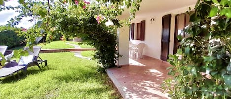 Un espace patio doté d’un coin salon couvert, d’une pelouse et d’un chemin menant à un bâtiment à l’extérieur blanc et aux volets sombres.